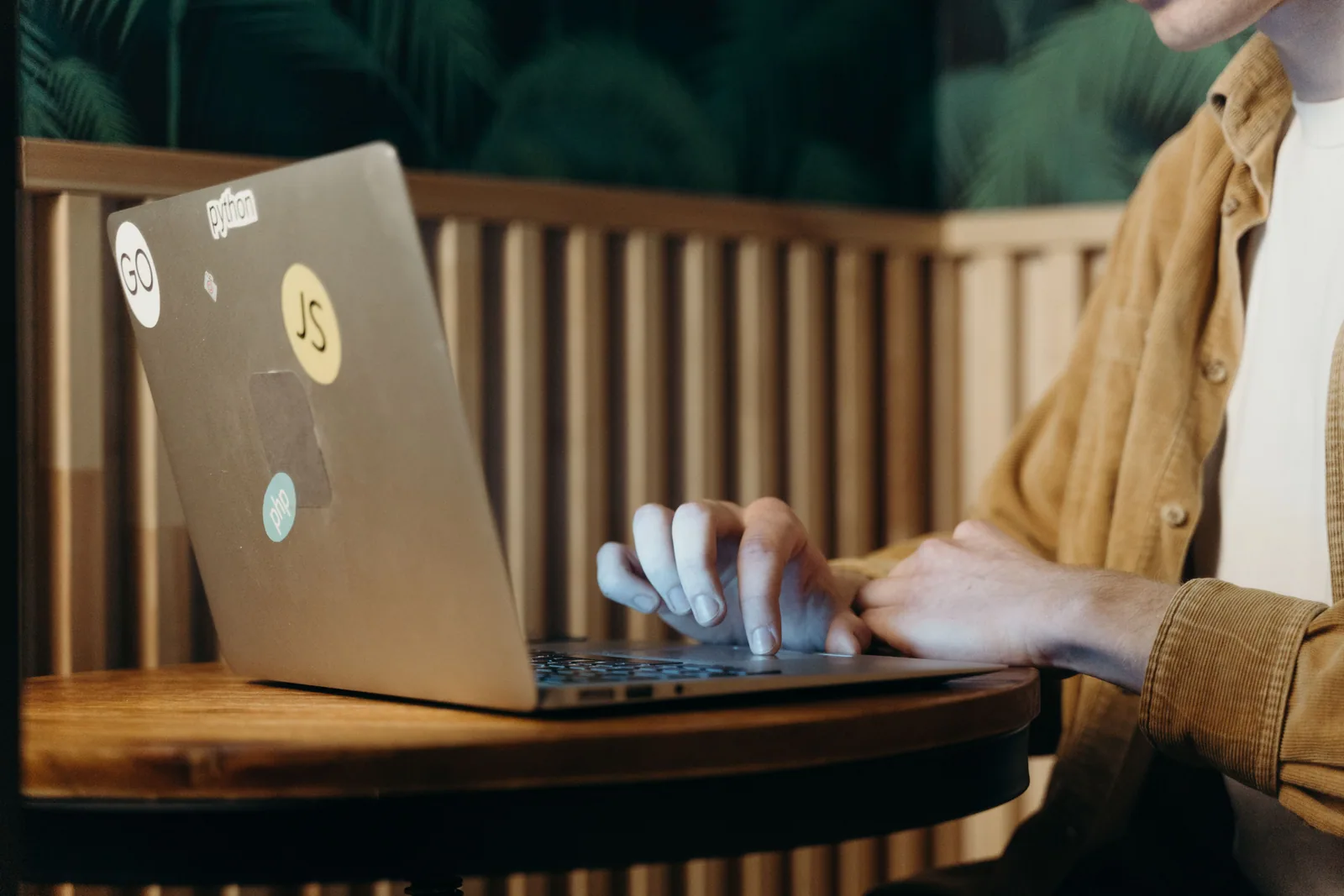 Hände tippen eine Frage in einen silbernen Laptop auf einem Holztisch in einem Café.