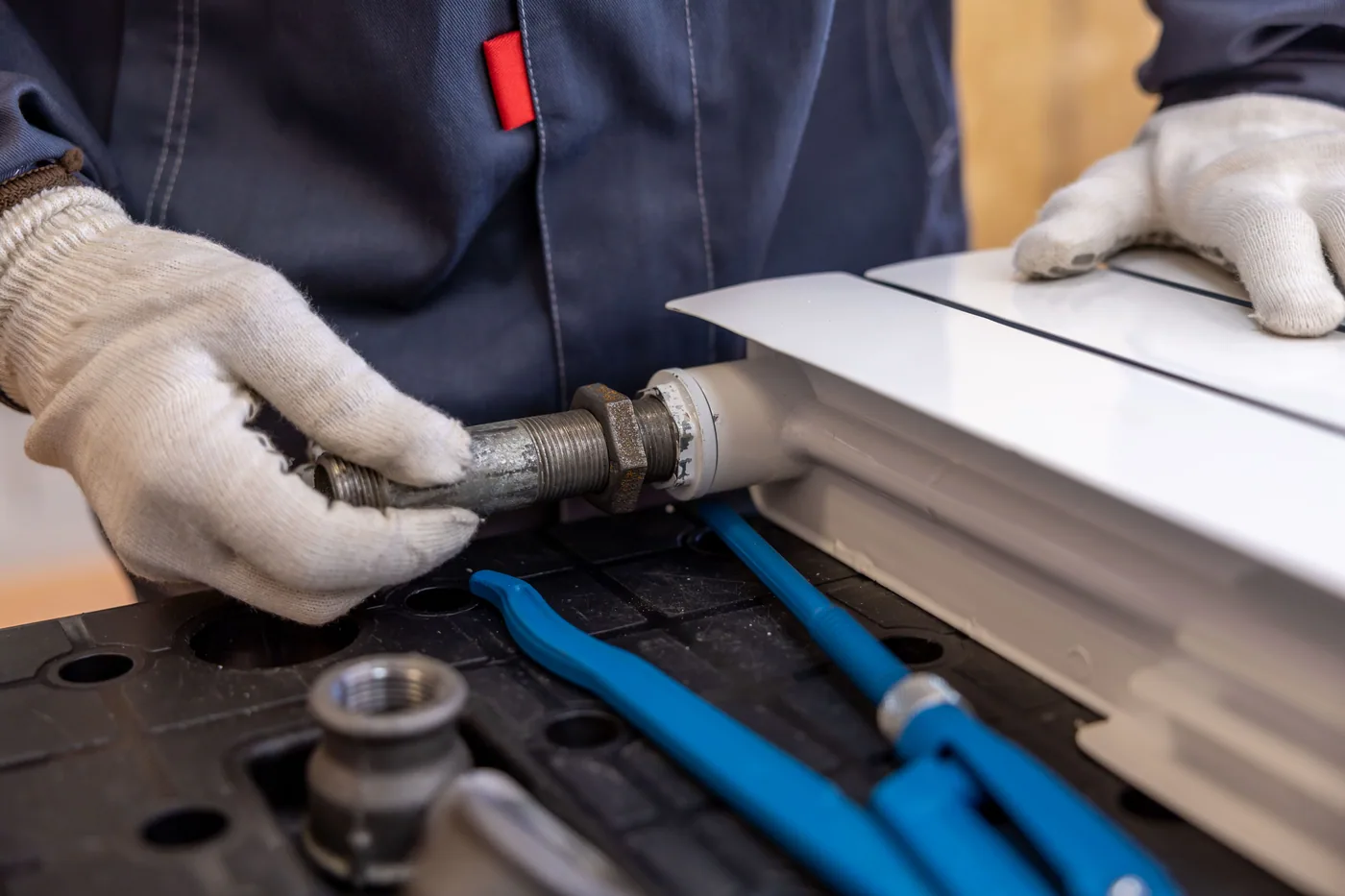 Hände in Arbeitshandschuhen verschrauben ein Heizungsrohr an einem Radiator — ein Sanitärbetrieb bei der Arbeit.