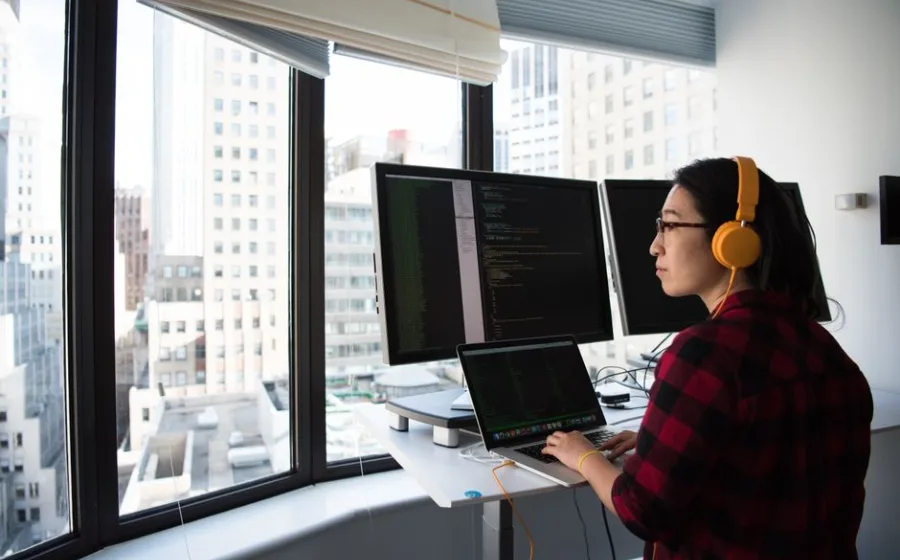 Entwicklerin am Stehpult vor zwei Bildschirmen mit Code, helles Büro mit Fensterfront