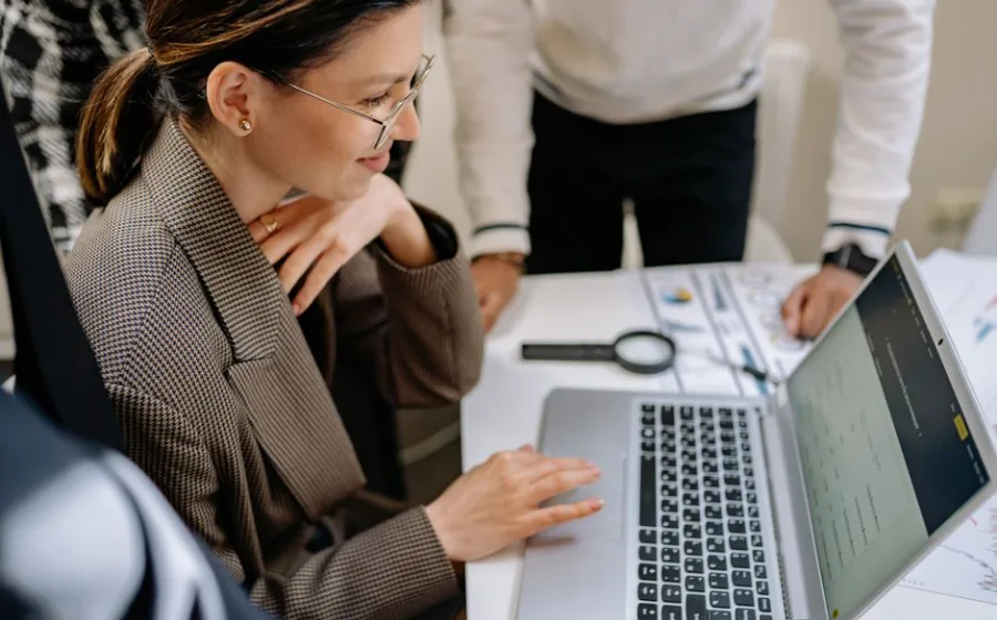 Mitarbeiterin lacht am Laptop, daneben Auswertungen und Kolleginnen — Marketing- und Office-Alltag in der Agentur